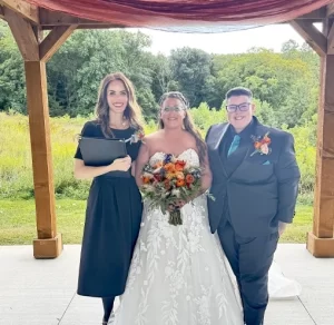 Indianapolis Wedding Officiant in black holding a book next to two women one in a white dress holding flowers and the other in a grey suit with a blue tie.
