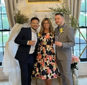 Weddings by Bretta Officiant in a flower dress with two men one in a grey suite and one in a black suit holding champagne glasses