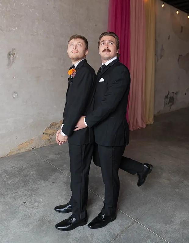 Two Grooms at Ivory Foundry Indianapolis Indiana — Wedding Coordination by Weddings by Bretta Two grooms in black tuxedos posing together in front of colorful draped backdrop at Ivory Foundry Indianapolis Indiana — wedding coordination by Weddings by Bretta
